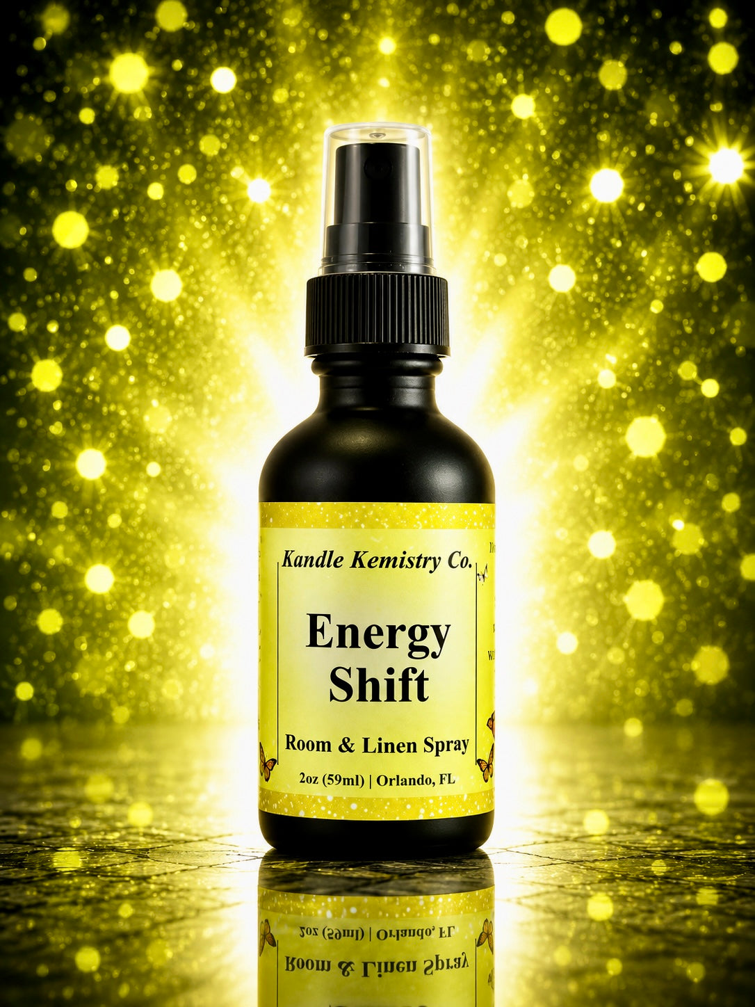 A bottle of Energy Shift Room & Linen Spray by Kandle Kemistry Co. The product is in a black glass bottle with a black spray nozzle and a pale yellow label, set against a sparkling gold bokeh background with a reflective surface below.