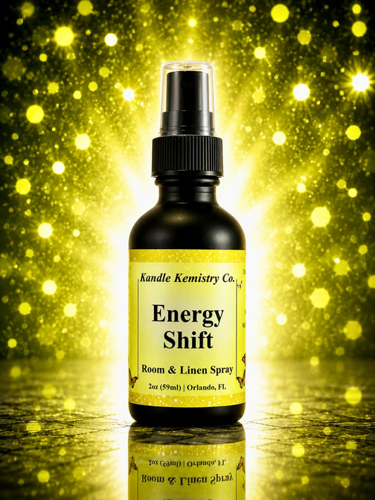 A bottle of Energy Shift Room & Linen Spray by Kandle Kemistry Co. The product is in a black glass bottle with a black spray nozzle and a pale yellow label, set against a sparkling gold bokeh background with a reflective surface below.