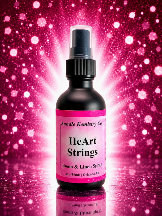 A bottle of HeArt Strings Room & Linen Spray by Kandle Kemistry Co. The product is in a matte black bottle with a pink label and a clear spray cap, set against a vibrant pink glittery background with a bright light burst behind it.