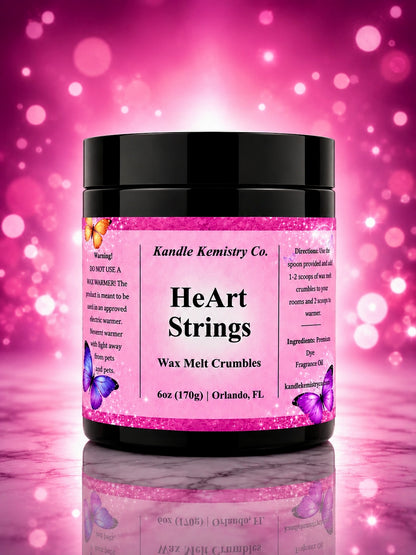 A jar of Kandle Kemistry Co. HeArt Strings Wax Melt Crumbles, featuring a black lid and a pink glittery label with purple butterflies, set against a pink bokeh background with a marble surface reflection.