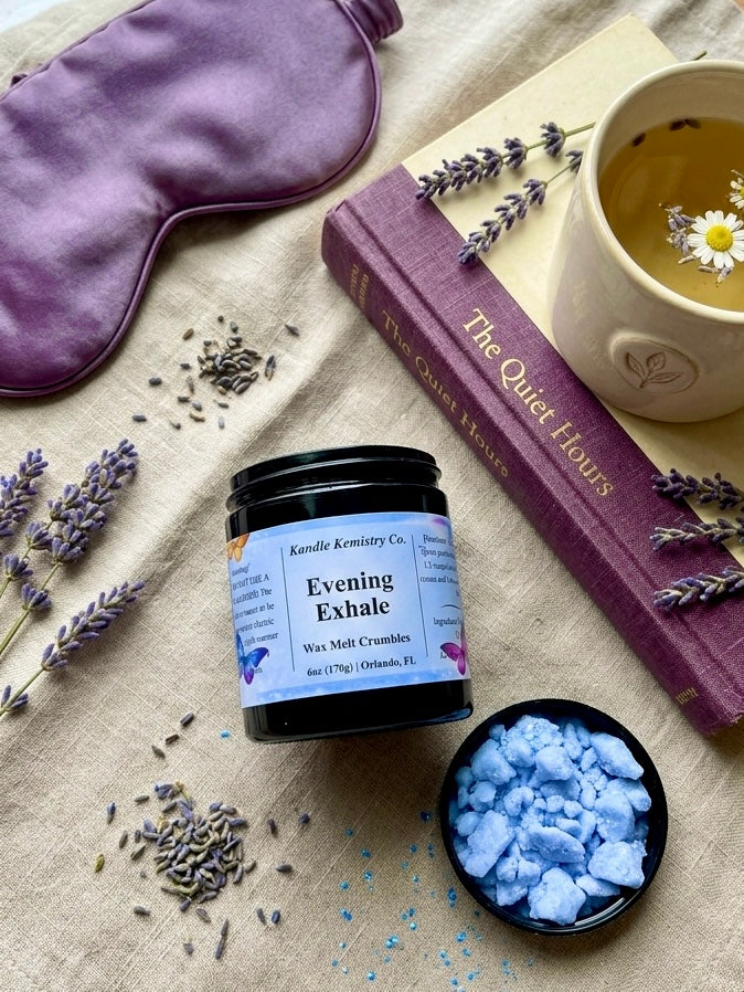 Candle with 'Evening Exhale' wax melts label, lavender flowers, a book titled 'The Quiet Hours', and a cup of tea on a textured surface.