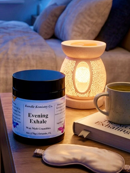 Jar of 'Evening Exhale' wax melt crumbles with a lit aromatherapy lamp, mug, and sleep mask on a table.