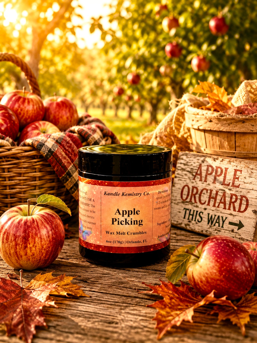 A jar of Kandle Kemistry Co. Apple Picking Wax Melt Crumbles sits on a rustic wooden table surrounded by red apples, autumn leaves, wicker baskets, and a wooden sign reading 'Apple Orchard This Way,' with a blurred apple orchard in the background.