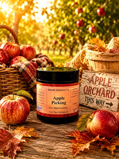 A jar of Kandle Kemistry Co. Apple Picking Wax Melt Crumbles sits on a rustic wooden table surrounded by red apples, autumn leaves, wicker baskets, and a wooden sign reading 'Apple Orchard This Way,' with a blurred apple orchard in the background.