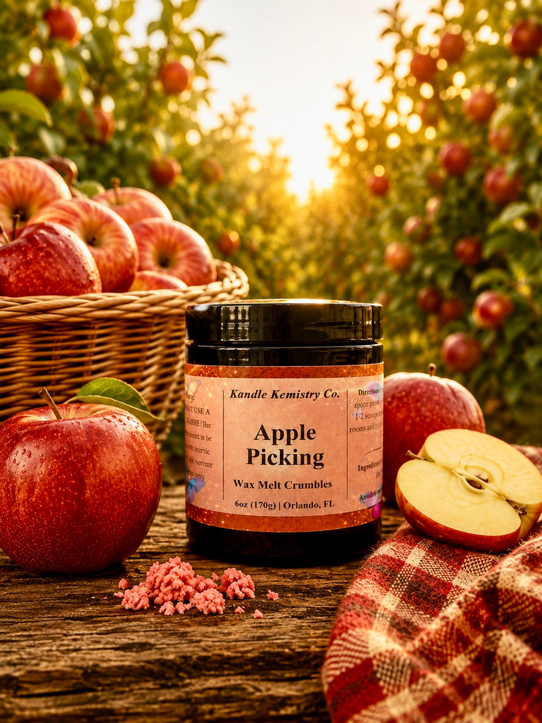 A jar of Kandle Kemistry Co. Apple Picking Wax Melt Crumbles sits on a rustic wooden table surrounded by fresh red apples, a basket of apples, and a plaid blanket, with an apple orchard in the background at sunset.