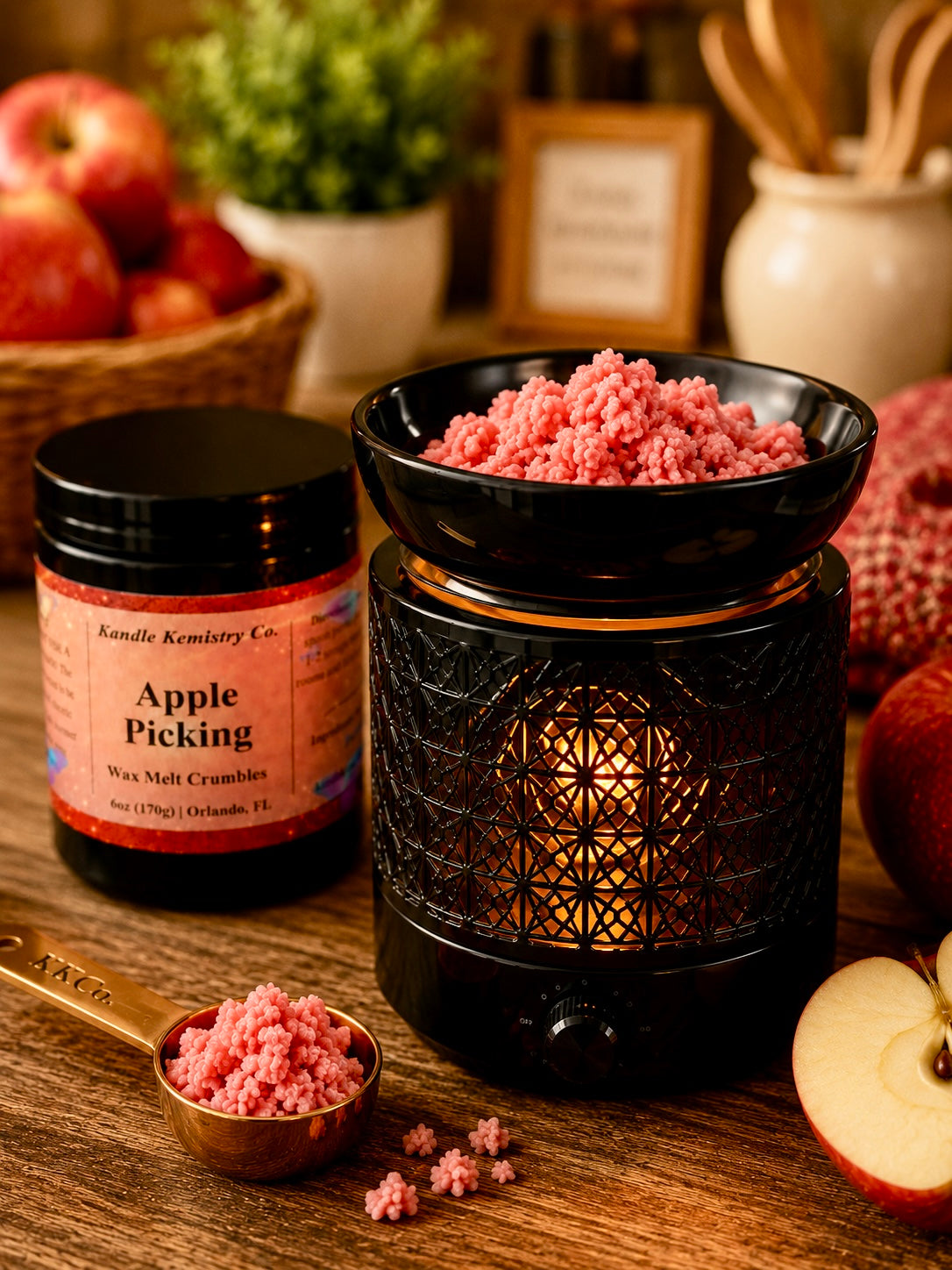A jar of Kandle Kemistry Co. Apple Picking wax melt crumbs sits beside a black decorative warmer filled with pink wax melt pieces, with apples and a wooden tabletop in a styled setting.
