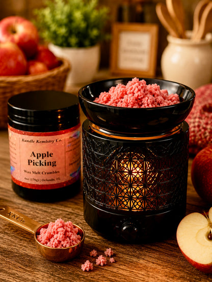 A jar of Kandle Kemistry Co. Apple Picking wax melt crumbs sits beside a black decorative warmer filled with pink wax melt pieces, with apples and a wooden tabletop in a styled setting.