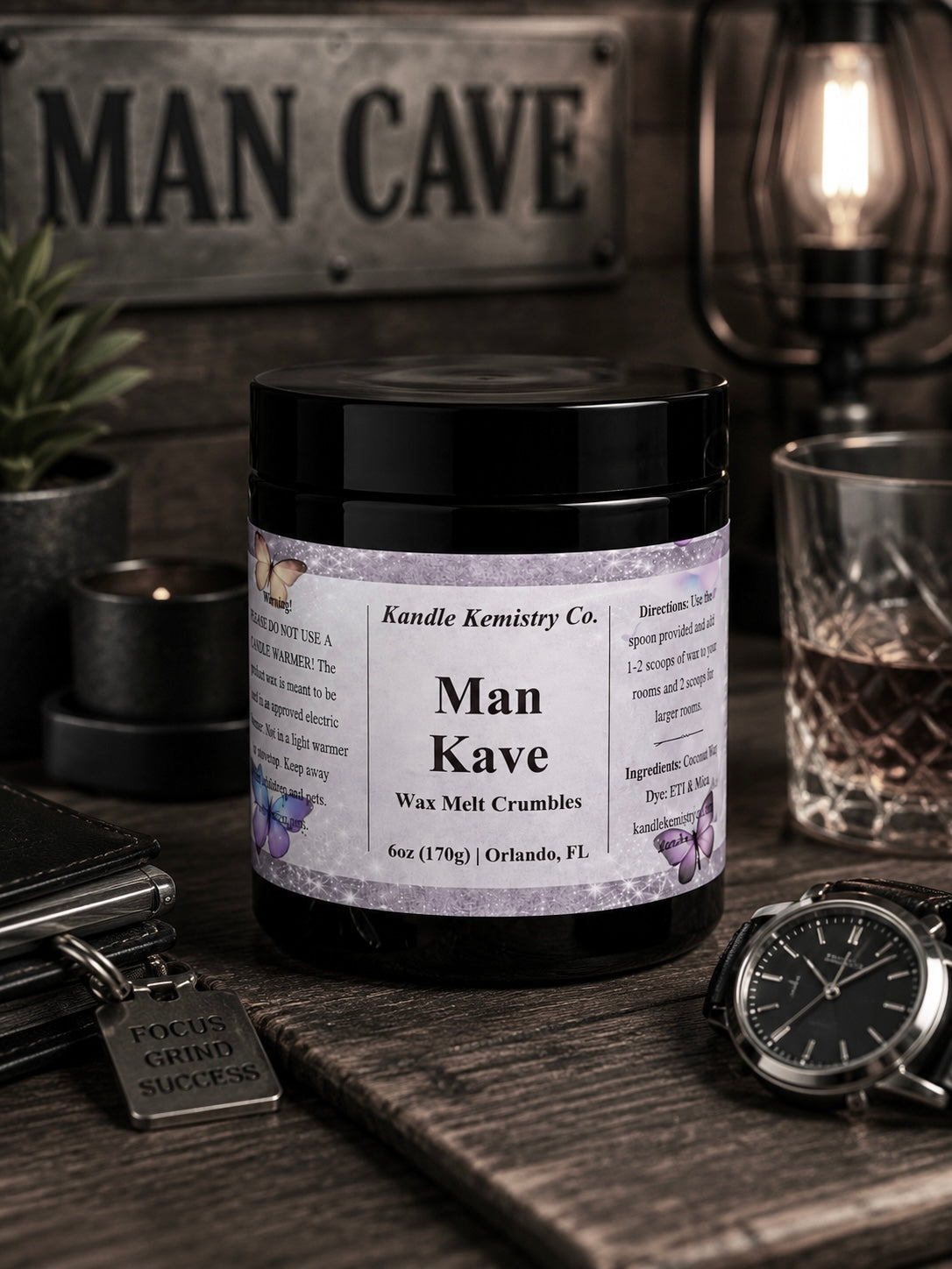 A black jar labeled 'Man Kave wax melt crumbles' by Kandle Kemistry Co., styled on a rustic wooden surface with a watch, wallet, glass, and a sign reading 'MAN CAVE' in the background.