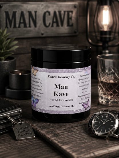 A black jar labeled 'Man Kave wax melt crumbles' by Kandle Kemistry Co., styled on a rustic wooden surface with a watch, wallet, glass, and a sign reading 'MAN CAVE' in the background.