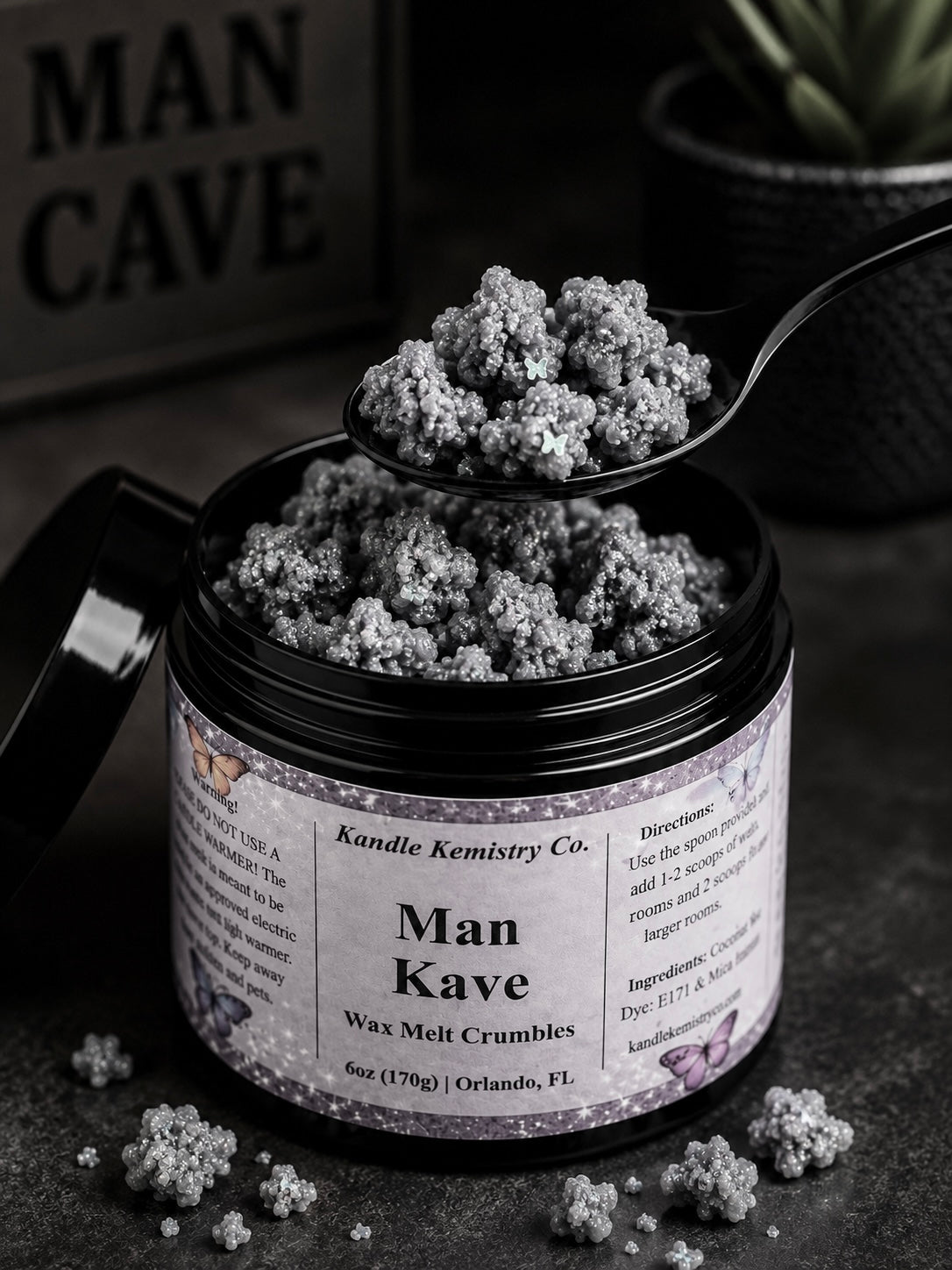 An open black jar of Man Kave Wax Melt Crumbles by Kandle Kemistry Co. sits on a dark surface with a spoon holding some of the crumbly product above the jar.