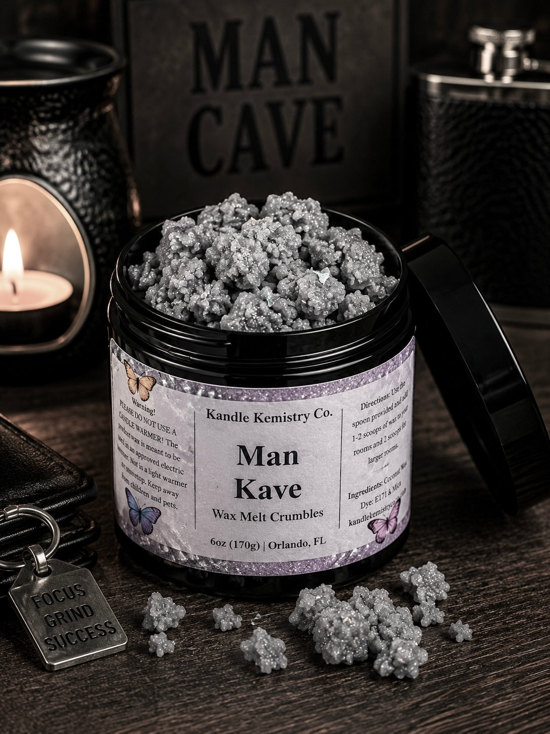 An open black jar of Man Kave Wax Melt Crumbles by Kandle Kemistry Co. sits on a dark wooden surface with a few crumbles spilled in front.