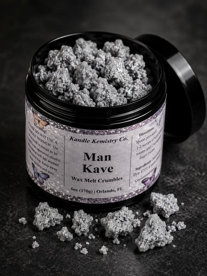 An open black jar of Man Kave Wax Melt Crumbles by Kandle Kemistry Co. on a dark textured surface, with some of the gray-white crumb product scattered in front and the lid resting behind the jar.