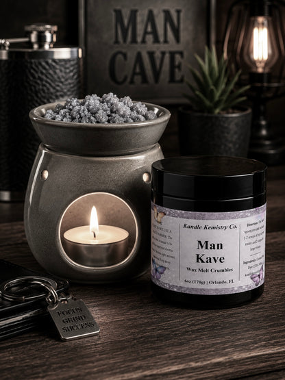 A black  jar labeled 'Man Kave' wax melt crumbles by Kandle Kemistry Co. sits next to a gray wax warmer with a lit tealight candle, keys, and a 'MAN CAVE' sign in a moody, dark-themed room setting.