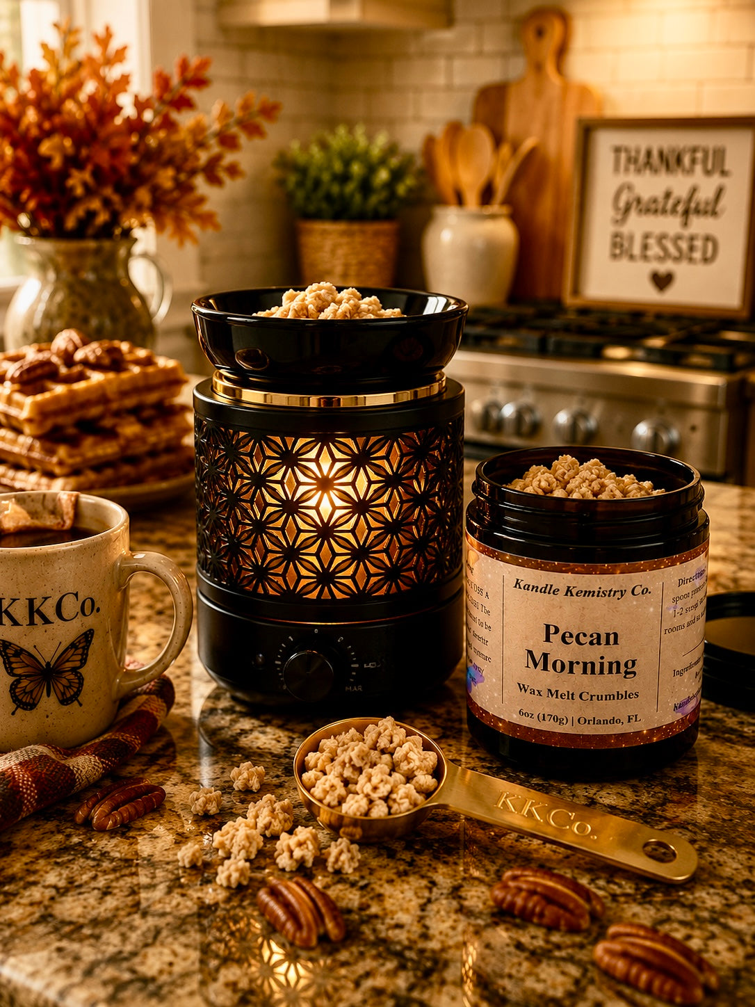 A kitchen countertop scene showing a black wax melt container with gold geometric cutout detailing, a jar labeled 'Pecan Morning Wax Melt Crumbles by Kandle Kemistry Co., a mug with KKCo.