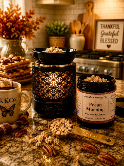 A kitchen countertop scene showing a black wax melt container with gold geometric cutout detailing, a jar labeled 'Pecan Morning Wax Melt Crumbles by Kandle Kemistry Co., a mug with KKCo.