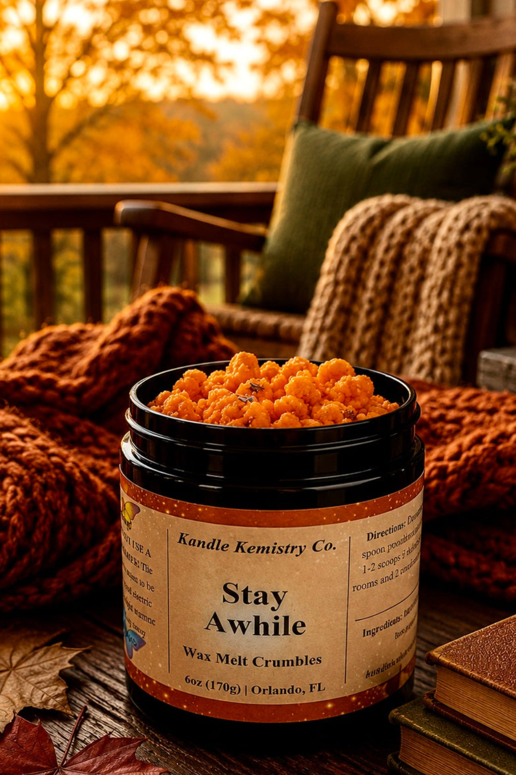 A candle jar labeled 'Stay Awhile Wax Melt Crumbles' by Kandle Kemistry Co. sits on a rustic wooden table on a porch, surrounded by a chunky orange knit blanket, autumn leaves, books, and a warm sunset view in the background.