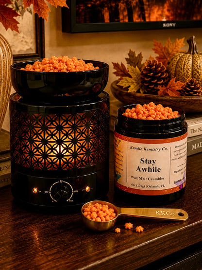 A jar of Kandle Kemistry Co. Stay Awhile Wax Melts Crumbles sits on a dark wooden table next to a black wax melt warmer filled with orange wax melts.