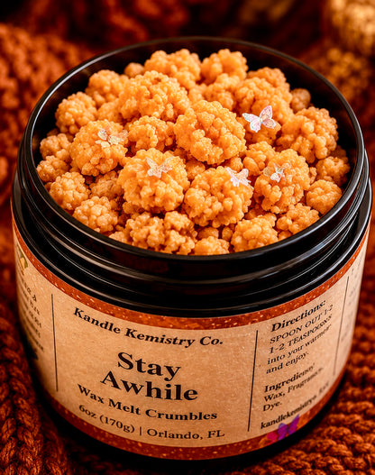An open black jar filled with orange-brown wax melt crumbles, labeled 'Stay Awhile' by Kandle Kemistry Co., set against a warm, textured brown knit background.