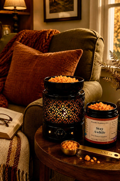 A cozy living room scene with a black decorative electric wax melt warmer filled with orange-yellow wax melt crumbles, alongside a jar labeled 'Stay Awhile Wax melt crumbles' and a measuring spoon on a wooden side table. The background features a warm-lit armchair, throw blanket, bookshelf, and window with autumn foliage.