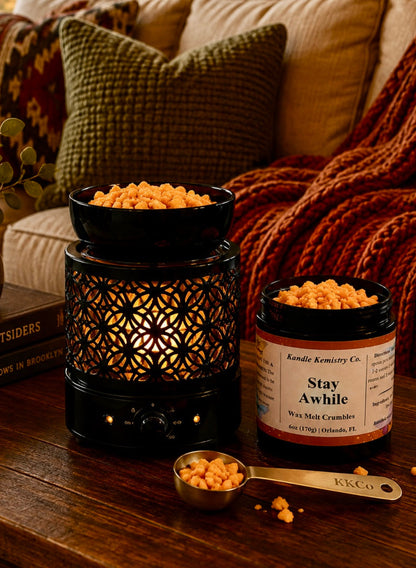 A cozy living room scene with a black decorative candle warmer and an open jar of Kandle Kemistry Co. Stay Awhile wax melt crumbles on a wooden table, with a metal scoop in front and a knitted orange-brown throw blanket draped over the sofa in the background.