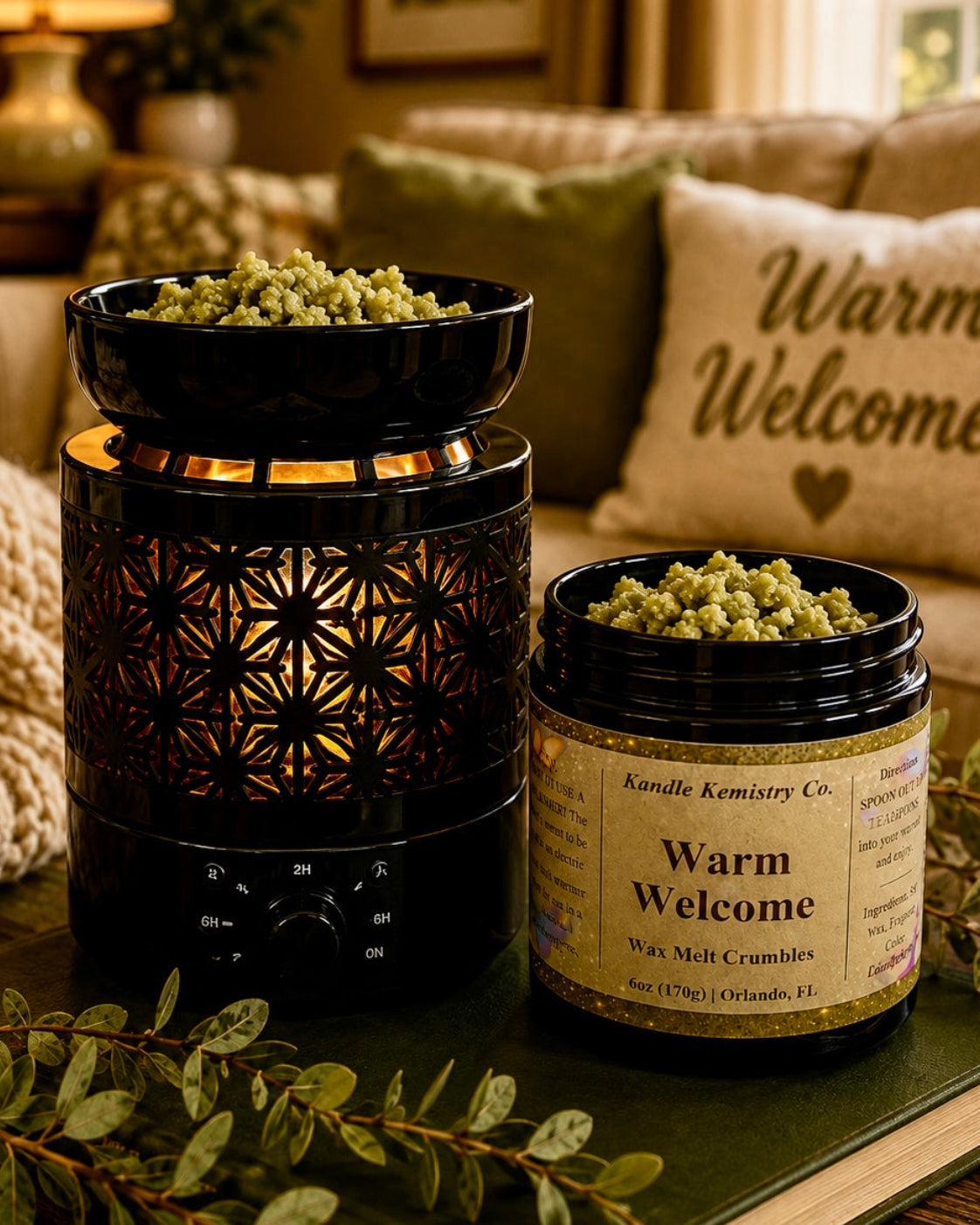 A jar of Kandle Kemistry Co. 'Warm Welcome' wax melt crumbles sits next to a black electric wax warmer with a matching black bowl, both filled with the greenish-yellow wax melts, styled on a book in a cozy living room setting.