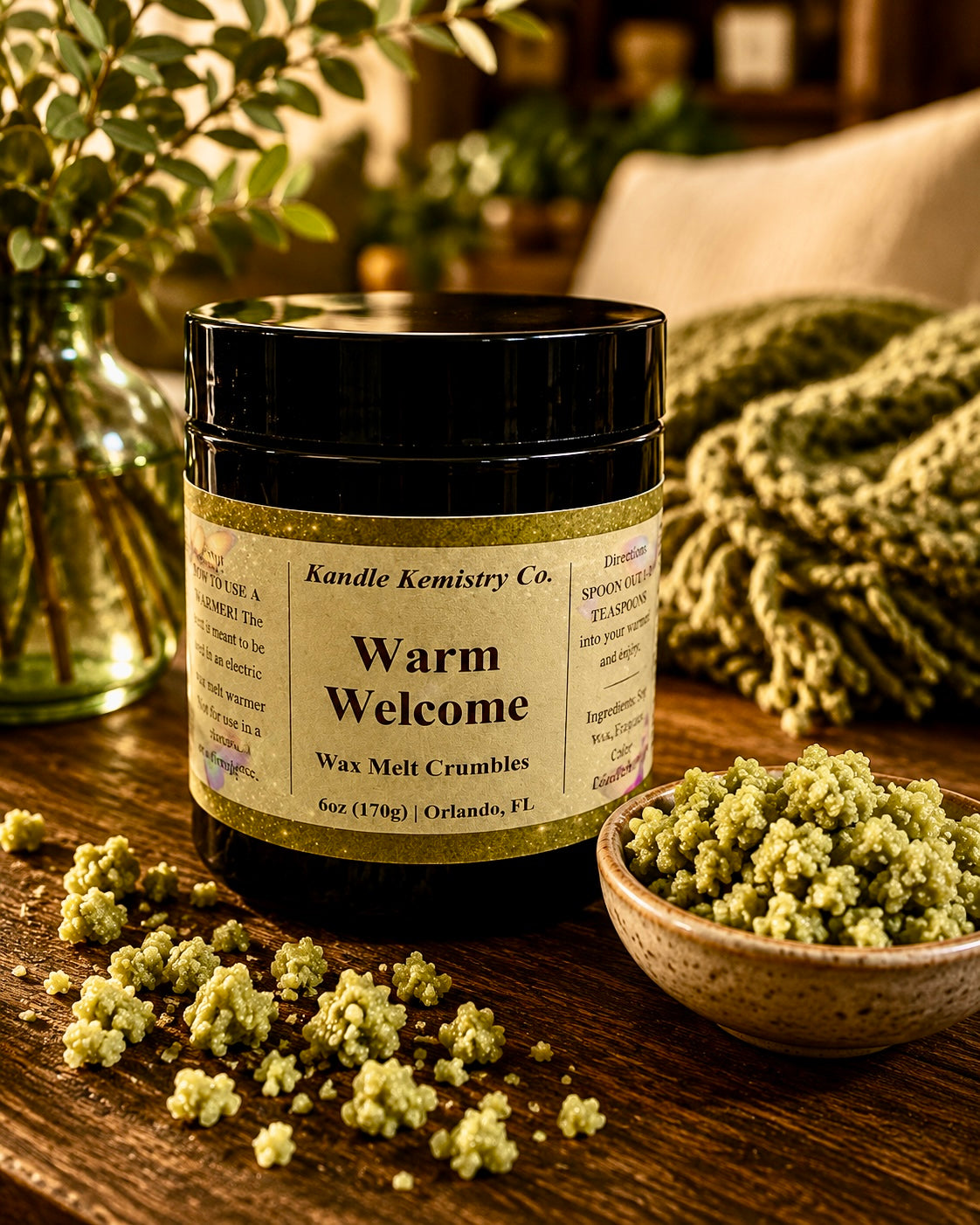 A black jar of Kandle Kemistry Co. 'Warm Welcome' wax melt crumbles sits on a rustic wooden table, surrounded by spilled wax crumbles, a small bowl of the greenish-yellow wax melts, and a cozy, warm-toned home background with a blanket and vase of greenery.