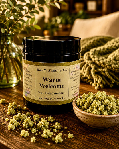 A black jar of Kandle Kemistry Co. 'Warm Welcome' wax melt crumbles sits on a rustic wooden table, surrounded by spilled wax crumbles, a small bowl of the greenish-yellow wax melts, and a cozy, warm-toned home background with a blanket and vase of greenery.
