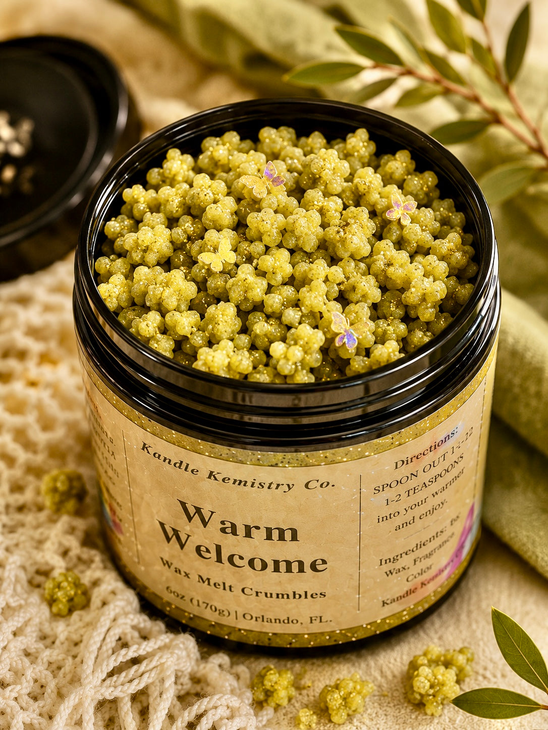 An open black jar filled with olive green wax melt crumbles, labeled 'Warm Welcome' by Kandle Kemistry Co., styled on a textured cream fabric with scattered wax pieces and soft botanical props.