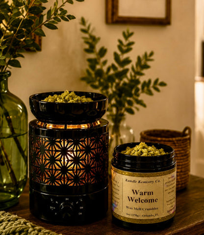 A styled home interior scene showing two wax melt products on a wooden table: a black metal wax melt warmer and green wax melt crumbles on top, and a black jar labeled 'Warm Welcome Wax melt crumbles' by Kandle Kemistry Co. Decorative greenery in glass vases and a knit blanket are visible around the products.